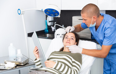 Professional cosmetologist examining female client face before procedure in esthetic clinic. Woman looking at mirror