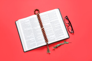 Open Holy Bible, eyeglasses and prayer beads on red background