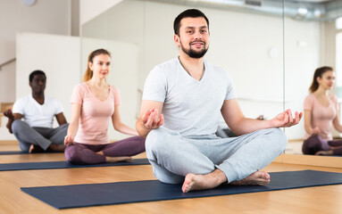 Young smiling man sitting in lotus pose on mat in fitness room, doing yoga meditation with group of adult people..
