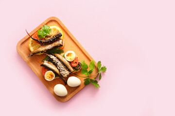 Wooden board of tasty sandwiches with canned smoked sprats on pink background