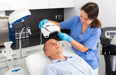 Focused female cosmetologist putting on spectrum LED mask preparing man for face and neck skin...