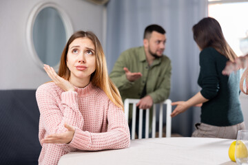 Sad woman sitting at table at home while man and other woman standing behind and quarreling.