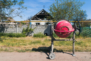 Countryside. A repair tool (concrete mixer) against the background of a house damaged by shelling....