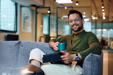 Happy businessman using touchpad while having cup of coffee at work and looking at camera. - obrazy, fototapety, plakaty