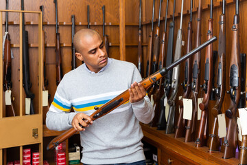 Portrait of focused Latino inspecting sporting shotgun before buying in gunsmith shop