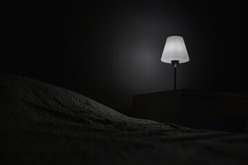 A bedside table and a burning lamp stands on it near the bed in a dark room. Hotel number. Place for text.