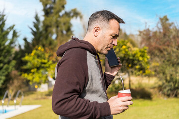 Fototapeta premium Adult man listening to messages on his phone and drinking mate infusion while walking in his garden. Home office concept.