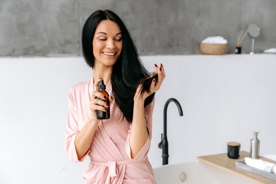 Haircare concept. Split ends repair treatment. Female beauty routine. Happy brunette caucasian woman applying essential oil spray, fluid, on her long hair, sits in a bathroom, looks at camera, smiles