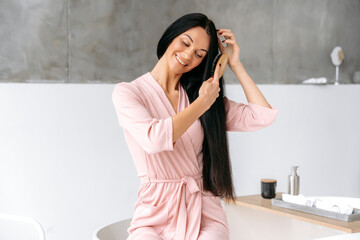 Female beauty routine. Combing hair. Beautiful caucasian brunette woman, sits in a stylish modern bathroom, in a bathrobe, combs her long hair with a wooden comb, smiles