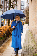 Rainy weather concept. A woman in a blue coat with a blue umbrella on a city street. Woman forty plus age.