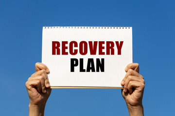 Recovery plan text on notebook paper held by 2 hands with isolated blue sky background. This message can be used as business concept about recovery plan.