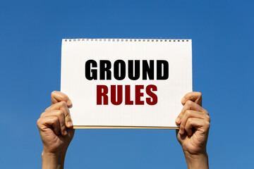 Ground rules text on notebook paper held by 2 hands with isolated blue sky background. This message can be used as business concept about ground rules.