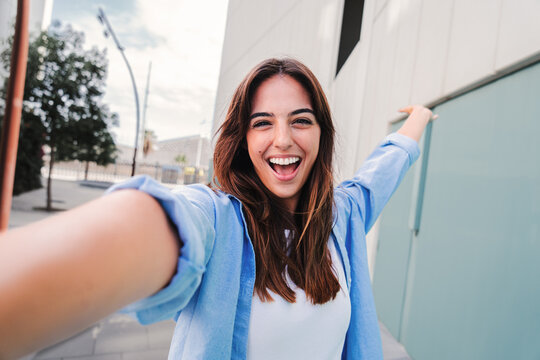 Happy young caucasian student lady looking at camera and taking a selfie portrait having fun, standing outside. Front view of laughing woman shooting a photo for social media at the university campus