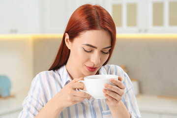 Beautiful woman with red dyed hair enjoying cup of drink in kitchen