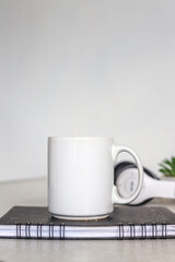 White coffee mug mockup on the book with blurry headphones in the background. Empty mug mock up for design promotion.