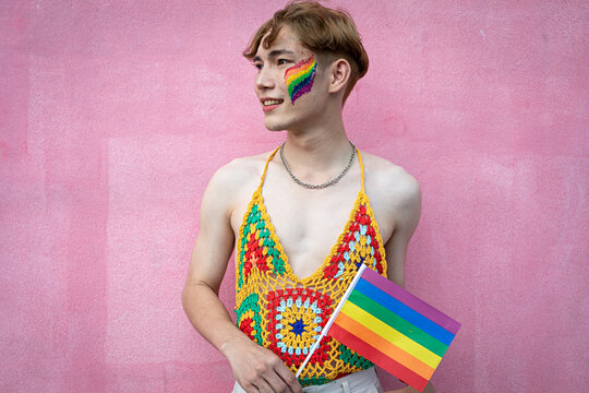 Portrait of Asian gay man with pride, holding LGBT Rainbow flag against pink background.