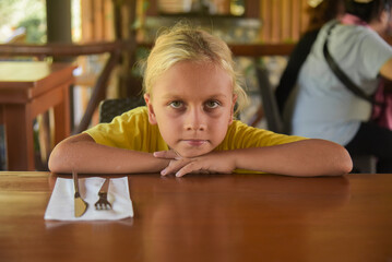 Fototapeta na wymiar An american blonde boy waiting for a dinner in restaurant. Portrait of child with long blonde hair and a yellow T-shirt sitting in a cafe. Bad appetite and digestive problems on a diet