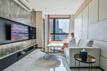 Tourist woman in a white bathrobe sits on a couch nearly window, watching TV in a modern room overlooking the city and skyscrapers. Holidays in a luxury hotel, travel and vacation.