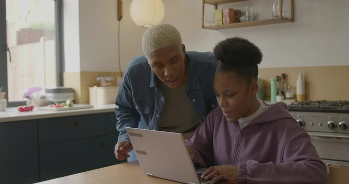 Mother helping Teenager Daughter with Homework on Laptop in Kitchen