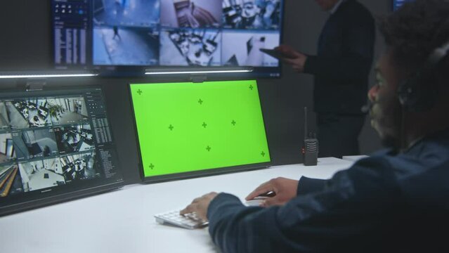 African American male operator works in security control center. Big digital screen and computer monitors with surveillance CCTV cameras video footage. Worker with tablet on background. Green screen.