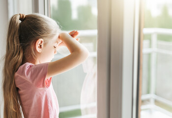 Sad little girl looking through  window at home.