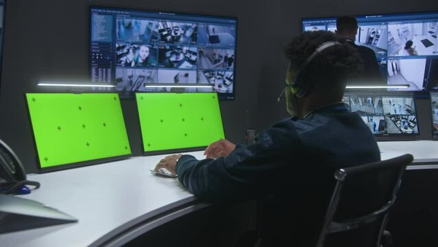 Mixed race officers, operators control security cameras. Employees work in monitoring center. Big digital screens showing surveillance CCTV cameras video footage. Computer monitors with green screen.
