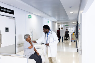 African american male doctor with tablet talking to diverse senior female patient in busy corridor