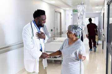 Biracial senior female patient with drip shaking hand of happy diverse male doctor in corridor