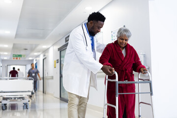 Diverse male doctor helping senior female patient use walking frame in hospital corridor, copy space