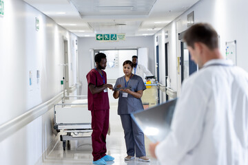 Diverse male and female doctor in scrubs looking at tablet talking in busy hospital corridor