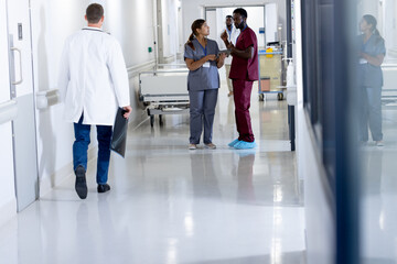 Diverse male and female doctor in scrubs with tablet, talking in busy hospital corridor, copy space