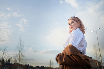 Beautiful girl with red hair in short skirt, white shirt and raincoat in village or small town. Tall young slender woman and houses and sky with clouds on background on an autumn, spring or summer day