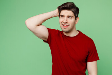 Young sad mistaken puzzled confused brunet caucasian man he wears red t-shirt casual clothes look aside scratch hold head isolated on plain pastel green background studio portrait. Lifestyle concept.