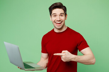 Young smiling happy smart IT man he wearing red t-shirt casual clothes hold use work on laptop pc...