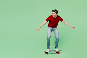 Full body side view happy fun cheerful young man he wears red t-shirt casual clothes driving skateboard pennyboard isolated on plain pastel light green background studio portrait. Lifestyle concept.
