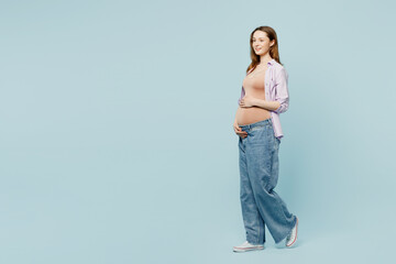 Full body sideways profile fun young pregnant future mom woman with belly tummy with baby wear casual clothes stroking stomach walk go isolated on plain blue background. Maternity pregnancy concept.