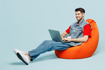Full body fun young IT man wear denim vest red t-shirt casual clothes sit in bag chair hold use work on laptop pc computer isolated on plain pastel light blue cyan background studio Lifestyle concept