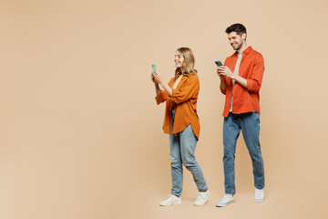 Full body sideways fun young couple two friends family man woman wear casual clothes hold in hand use mobile cell phone together isolated on pastel plain light beige color background studio portrait.