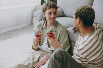 Young fun couple two gay men wear casual clothes together drink red wine talk on date sit on sofa...