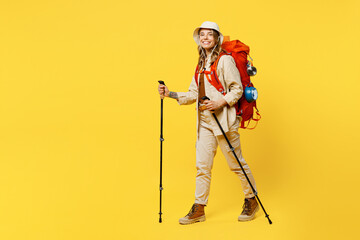 Full body sideways young woman carry bag with stuff mat hold trekking poles isolated on plain yellow background. Tourist leads active lifestyle walk on spare time Hiking trek rest travel trip concept.