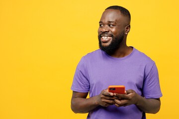 Young man of African American ethnicity wear casual clothes purple t-shirt hold in hand use mobile cell phone look aside on area isolated on plain yellow background studio portrait. Lifestyle concept.