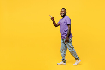 Full body young man of African American ethnicity he wear casual clothes purple t-shirt walk go stroll point index finger aside indicate on area copy space mock up isolated on plain yellow background.