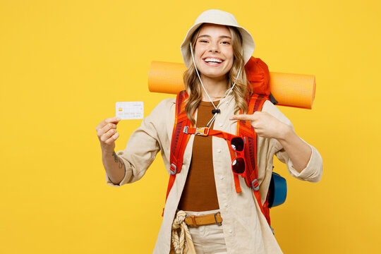 Young woman carry backpack with stuff mat hold point on credit bank card isolated on plain yellow background. Tourist leads active lifestyle walk on spare time. Hiking trek rest travel trip concept.