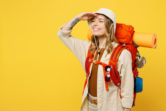 Young woman carry backpack with stuff mat hold hand at forehead look far away isolated on plain yellow background. Tourist leads active lifestyle walk spare time. Hiking trek rest travel trip concept.
