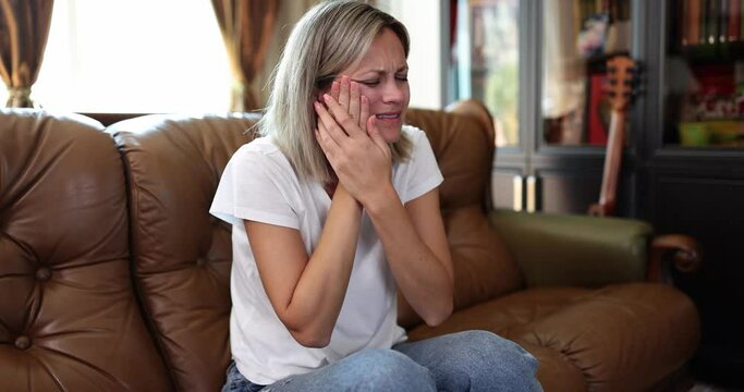Blonde woman grimaces feeling acute tooth ache putting hands on cheek. Female person suffers from pain sitting on soft sofa at home slow motion