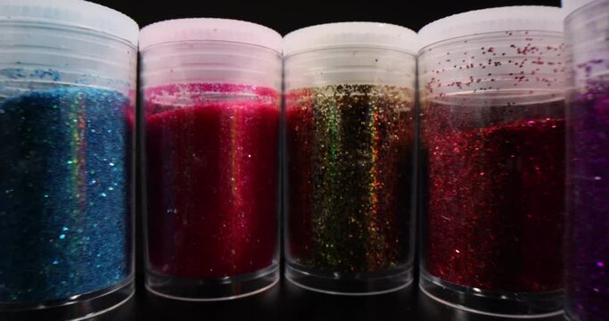 Sealed jars with colorful shiny glitter powder standing in curvy row on dark table. Bright decoration for makeup and nails on black background