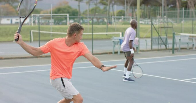 Two diverse male friends playing doubles returning ball on outdoor tennis court in slow motion