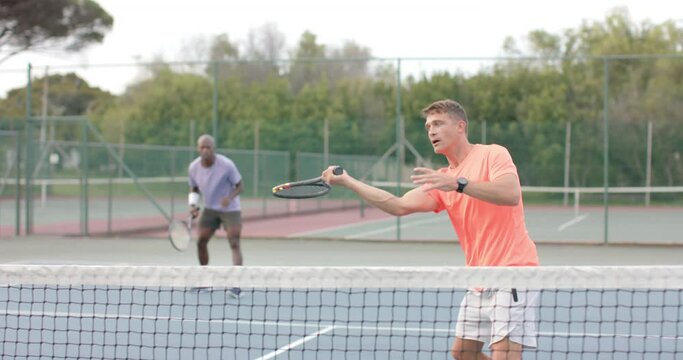 Two diverse male friends playing doubles returning ball over net at outdoor court in slow motion