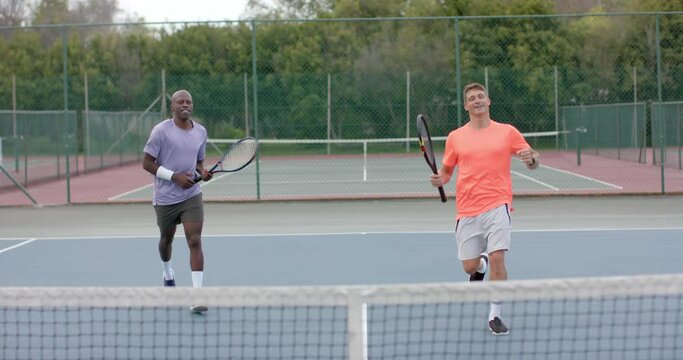 Two diverse male friends playing doubles celebrating winning point on outdoor court in slow motion