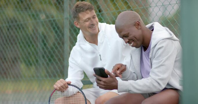 Happy diverse male friends looking at smartphone and laughing at tennis court, slow motion